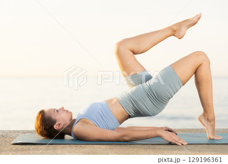 Sporty female wearing sportswear standing legs up in Salamba Sarvangasana exercise during yoga on seashore at sunrise 129196361