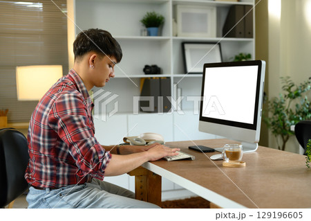 Side view of man working from home on desktop computer with coffee cup on wooden desk Side view of man working from home on desktop computer with coffee cup on wooden desk 129196605