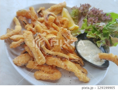 Golden, crispy fried squid calamari rings served on a dark slate plate with zesty lemon wedges and creamy garlic aioli. Golden, crispy fried squid calamari rings served on a dark slate plate with zesty lemon wedges and creamy garlic aioli. 129199905