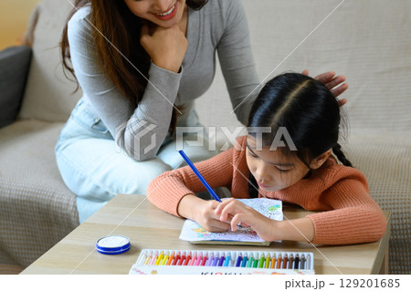 Happy mom supervising little girl doing coloring activity with colored pencils in a living room Happy mom supervising little girl doing coloring activity with colored pencils in a living room 129201685