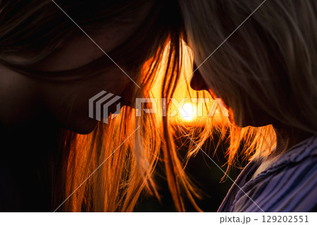 Tender moment between mother and teenage daughter touching foreheads during sunset with glowing backlight in their hair Tender moment between mother and teenage daughter touching foreheads during sunset with glowing backlight in their hair 129202551