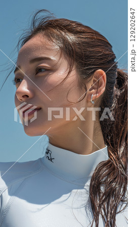 Young woman enjoys a sunny day at the beach while wearing a surf suit and looking off into the distance with a confident expression Young woman enjoys a sunny day at the beach while wearing a surf suit and looking off into the distance with a confident expression 129202647