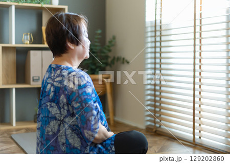 室内の床に座って目を閉じ、瞑想をする60代のシニア女性 129202860