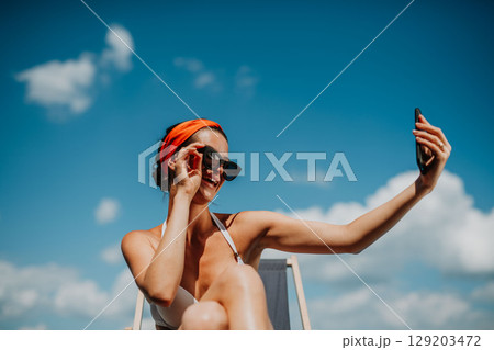 Young woman sitting by swimming pool outdoors in backyard garden, taking selfie. 129203472