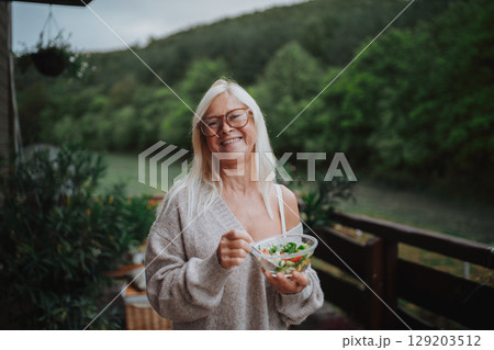 Senior woman standing terrace, eating healthy dinner outdoors. Senior woman standing terrace, eating healthy dinner outdoors. 129203512