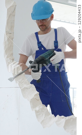 Male construction worker wearing blue overalls, protective helmet and white gloves, is demolishing white wall with rotary hammer drill, generating dust, close up horizontal view. Renovation concept Male construction worker wearing blue overalls, protective helmet and white gloves, is demolishing white wall with rotary hammer drill, generating dust, close up horizontal view. Renovation concept 129204433