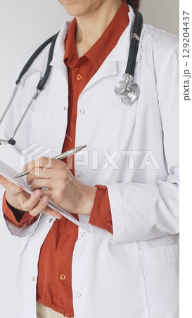 Close up of woman doctor wearing stethoscope and white coat over brick red blouse is standing and writing on a clipboard, representing medical expertise and patient care. Medicine concept 129204437