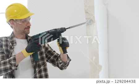 Portrait of a man construction worker wearing beige checkered shirt , yellow hard hat and protective gloves, is demolishing white wall with rotary hammer drill, generating dust. Renovation concept Portrait of a man construction worker wearing beige checkered shirt , yellow hard hat and protective gloves, is demolishing white wall with rotary hammer drill, generating dust. Renovation concept 129204438