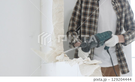 Unrecognizable male construction worker wearing beige checkered shirt and protective gloves, is demolishing white wall with rotary hammer drill, generating dust, close up view. Renovation concept 129204444