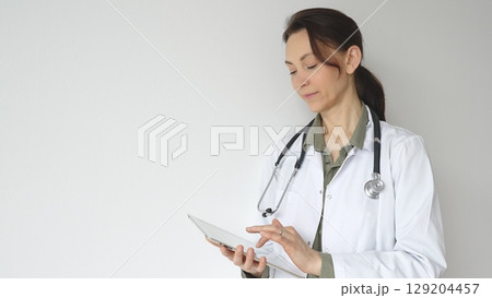 Headshot of a doctor woman wearing white medical coat over green blouse, stethoscope, is reviewing patient records on digital tablet, standing in medical workspace with natural lighting. Medicine Headshot of a doctor woman wearing white medical coat over green blouse, stethoscope, is reviewing patient records on digital tablet, standing in medical workspace with natural lighting. Medicine 129204457