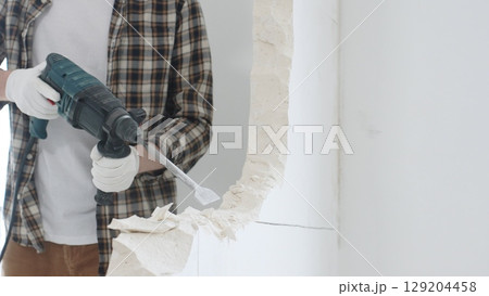Close up of unrecognizable male construction worker wearing beige checkered shirt and protective gloves, is demolishing white wall with rotary hammer drill, generating dust, close up view. Renovation Close up of unrecognizable male construction worker wearing beige checkered shirt and protective gloves, is demolishing white wall with rotary hammer drill, generating dust, close up view. Renovation 129204458