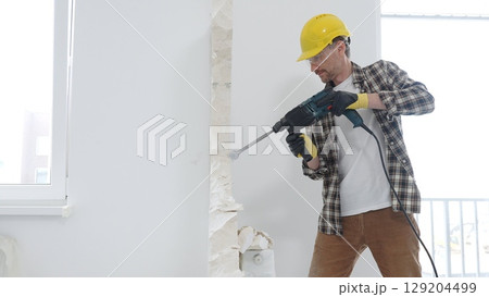 Man construction worker wearing beige checkered shirt , yellow hard hat and protective gloves, is demolishing white wall with rotary hammer drill, generating dust, medium long shot. Renovation concept 129204499