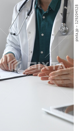 Doctor man wearing medical coat over green shirt, and stethoscope is gesturing with hands over white table while explaining diagnosis to patient. Medicine and health care concept 129204528