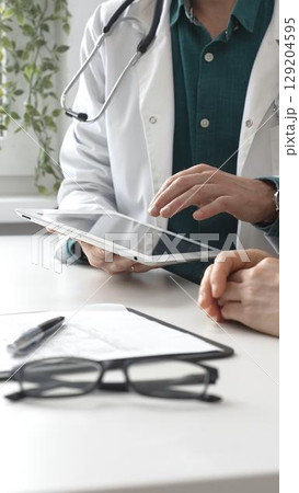 Male doctor in lab coat over a green shirt, and stethoscope gesturing while explaining diagnosis to patient using tablet in modern medical office. Medicine and health care concept 129204595