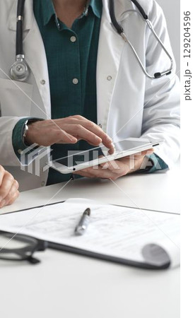Doctor man in lab coat over a green shirt, and stethoscope is using tablet while consulting patient in modern medical office. Medicine and health care concept 129204596