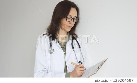 Doctor woman wearing stethoscope, black glasses and white coat over green blouse is standing and writing on a clipboard, representing medical expertise and patient care. Medicine and health care 129204597