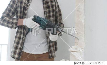 Unrecognizable male construction worker wearing beige checkered shirt and protective gloves, is demolishing white wall with rotary hammer drill, generating dust, close up view. Renovation concept 129204598