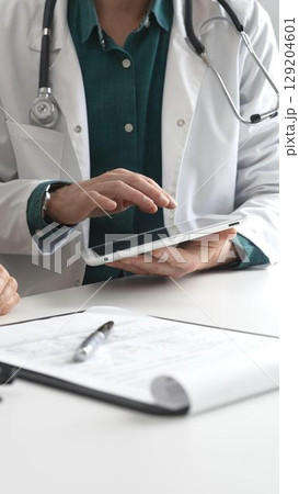 Doctor man in lab coat over a green shirt, and stethoscope is using tablet while consulting patient in modern medical office. Medicine and health care concept 129204601