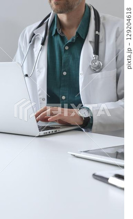 Doctor man wearing a white coat over green shirt, and stethoscope is typing on a laptop in a bright medical office, showcasing the intersection of healthcare and technology. Medicine and health care Doctor man wearing a white coat over green shirt, and stethoscope is typing on a laptop in a bright medical office, showcasing the intersection of healthcare and technology. Medicine and health care 129204618