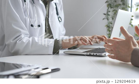 Doctor wearing lab coat and stethoscope typing on laptop keyboard and explaining something to patient during a medical consultation in hospital office. Medicine and health care concept Doctor wearing lab coat and stethoscope typing on laptop keyboard and explaining something to patient during a medical consultation in hospital office. Medicine and health care concept 129204619