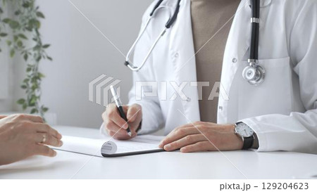 Doctor man listening to patient health concerns and writing a prescription on a clipboard during a consultation in a bright, modern clinic. Medicine and health care concept Doctor man listening to patient health concerns and writing a prescription on a clipboard during a consultation in a bright, modern clinic. Medicine and health care concept 129204623