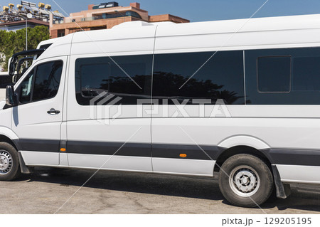 A large white van is parked on a sunny day, displaying a sleek design and tinted windows, surrounded by contemporary architecture in an urban area 129205195