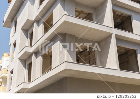A contemporary structure features sharp lines and large windows at different levels, reflecting modern architecture. The building is in the early stages of construction under a clear blue sky 129205252