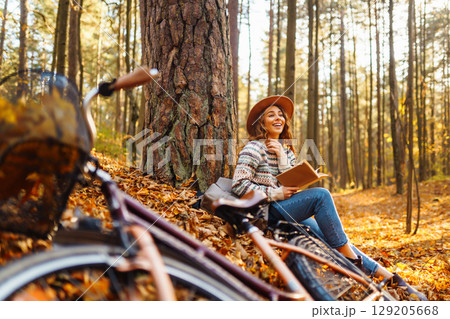 Beautiful young woman sitting on a fallen autumn leaves in a park, reading a book. 129205668