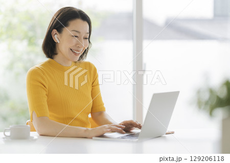 イヤホンをつけてノートパソコンに向かうアラフィフの女性 129206118