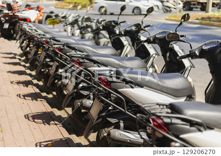A long line of scooters is parked neatly along the sidewalk. The bright, sunny day highlights the chrome and colors of the vehicles, contrasting with the busy street behind them 129206270