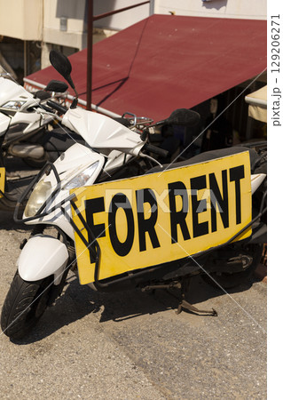 A clear sign indicating scooter rental services can be seen prominently displayed on a parked white scooter. The setting is vibrant, attracting tourists looking for transportation options 129206271