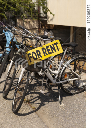 Multiple bicycles with baskets are lined up next to a wall, featuring a bright yellow sign indicating they are available for rent. The sun shines down, creating a vibrant atmosphere Multiple bicycles with baskets are lined up next to a wall, featuring a bright yellow sign indicating they are available for rent. The sun shines down, creating a vibrant atmosphere 129206272