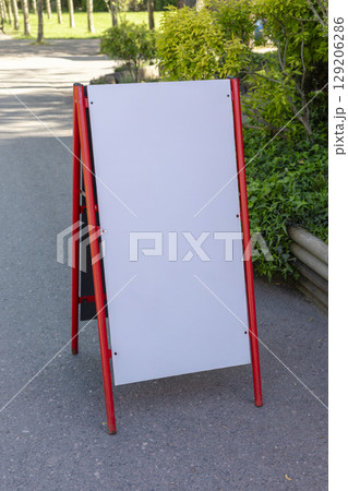 A blank outdoor sign stands on a pathway in a park filled with greenery. The sign is framed in bright red and awaits messages or advertisements. Sunlight illuminates the area. Mockup. 129206286