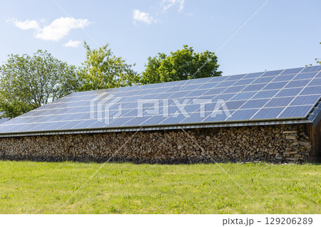 Solar panels cover the roof of a wooden shed, with neatly stacked firewood at its base. The bright green field and clear blue sky create a serene outdoor atmosphere Solar panels cover the roof of a wooden shed, with neatly stacked firewood at its base. The bright green field and clear blue sky create a serene outdoor atmosphere 129206289