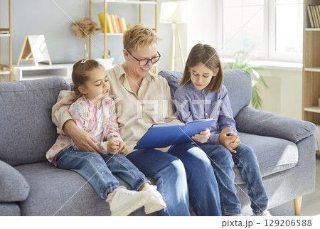 Smiling grandmother sitting on sofa and reading storybook to two children during family leisure time Smiling grandmother sitting on sofa and reading storybook to two children during family leisure time 129206588