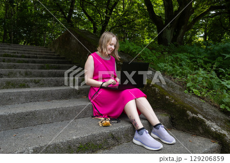 Young woman works on her laptop in a summer park, her pet agama lizard lounging peacefully beside her on a delicate leash Young woman works on her laptop in a summer park, her pet agama lizard lounging peacefully beside her on a delicate leash 129206859