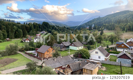 A serene, high-angle view of a small, traditional village nestled in a lush, green valley surrounded by forested mountains under a partly cloudy sky. Carpathian Mountains, Ukraine. A serene, high-angle view of a small, traditional village nestled in a lush, green valley surrounded by forested mountains under a partly cloudy sky. Carpathian Mountains, Ukraine. 129207019