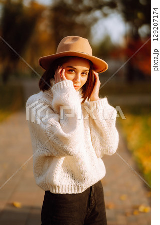 Autumn woman. Young cheerful woman in white sweater and hat walks in the nature park on a sunset. Autumn woman. Young cheerful woman in white sweater and hat walks in the nature park on a sunset. 129207174