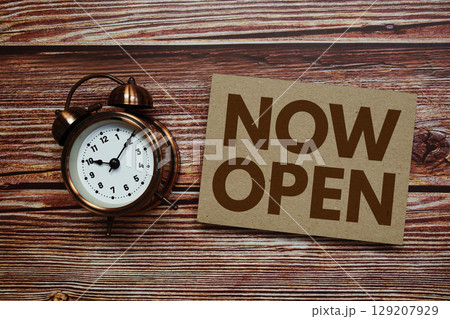 Now Open text with alarm clock on wooden background 129207929