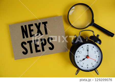 Next Steps text written on paper with magnifying glass and alarm clock on yellow background 129209076