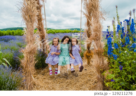 A warm breeze is playing in the long hair of the sisters who are swinging on a big swing in a lavender field 129211276