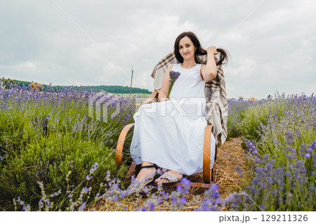 A young woman in a long white dress sits on a rocking chair in the middle of a lavender field 129211326