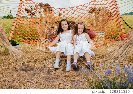 Two girls in white dresses are joyfully swinging on a red mesh hammock Two girls in white dresses are joyfully swinging on a red mesh hammock 129211530