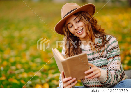 Young woman in hat sits on a plaid in autumn park and reads a book. Concept of nature, education. Young woman in hat sits on a plaid in autumn park and reads a book. Concept of nature, education. 129211759