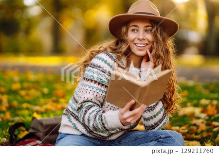 Young woman in hat sits on a plaid in autumn park and reads a book. Concept of nature, education. Young woman in hat sits on a plaid in autumn park and reads a book. Concept of nature, education. 129211760
