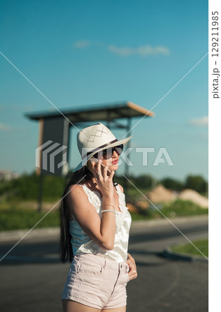 A young woman in a light white outfit is standing in the middle of nature and talking on the phone A young woman in a light white outfit is standing in the middle of nature and talking on the phone 129211985