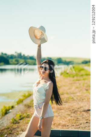 A young woman in a light-colored outfit raises her hat to the sky, as if greeting a sunny day A young woman in a light-colored outfit raises her hat to the sky, as if greeting a sunny day 129212004
