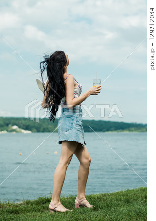 The girl in a hat has stopped by the lake, enjoying the drink in her cup The girl in a hat has stopped by the lake, enjoying the drink in her cup 129212034