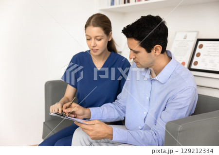 Young doctor in blue scrubs helping patient to fill application Young doctor in blue scrubs helping patient to fill application 129212334