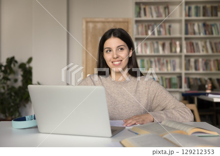 Happy girl student sitting at study desk smiling to aside 129212353